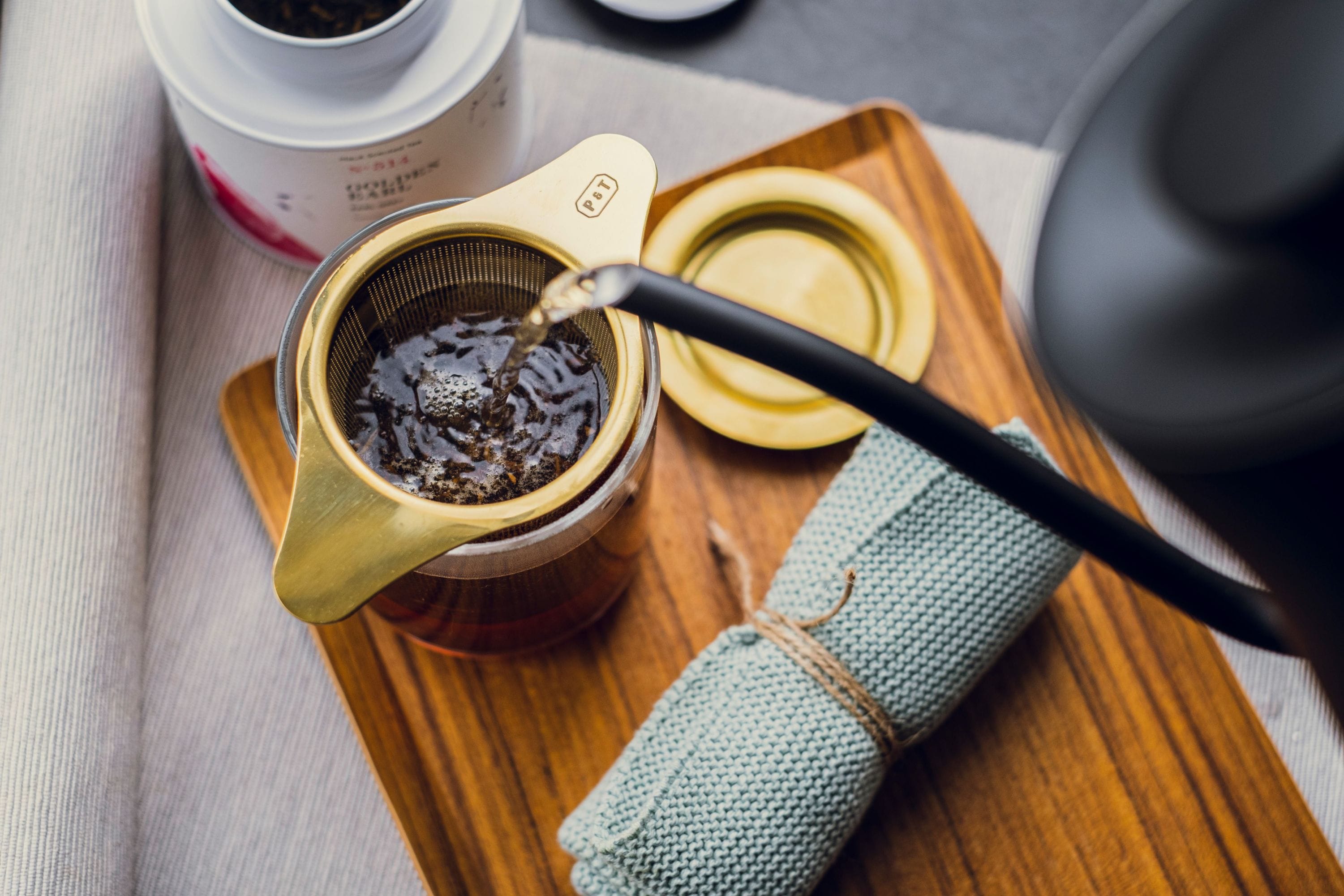 Heißes Wasser wird aus einem schwarzen Wasserkocher in einen Metallfilter mit Teeblättern gegossen, der über einem Glasbehälter steht