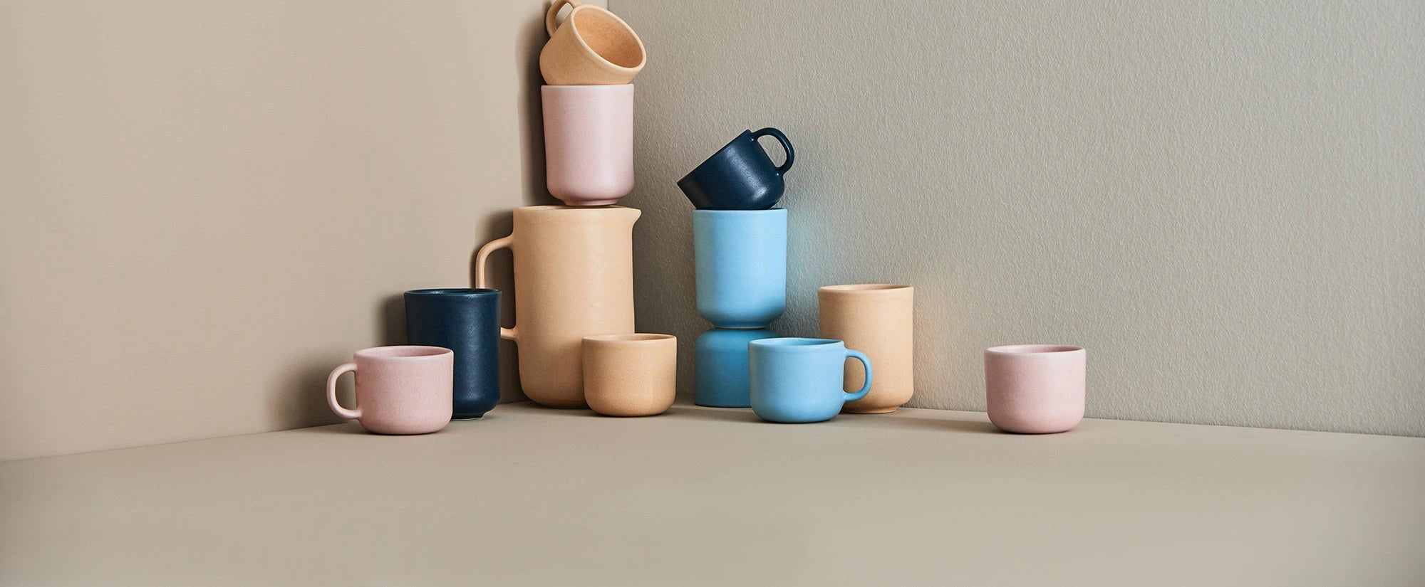 Keramiktassen und -kannen in Pastellfarben wie Rosa, Blau und Beige, gestapelt und gruppiert vor neutralem Hintergrund