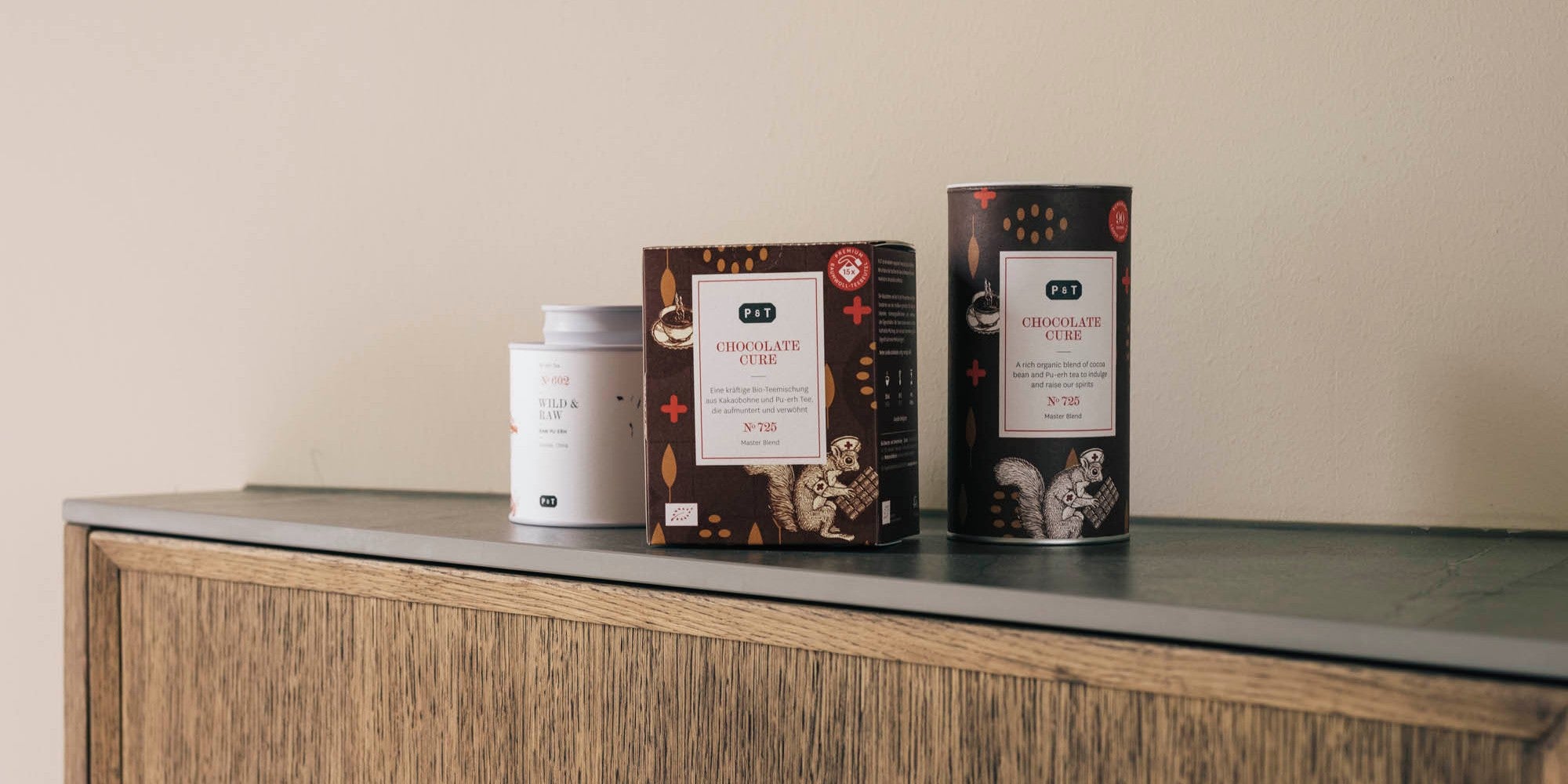 Drei Teeboxen auf Holzoberfläche: ein weißer zylindrischer Caddy, eine rechteckige Box und eine zylindrische Dose mit Tierillustrationen in Braun