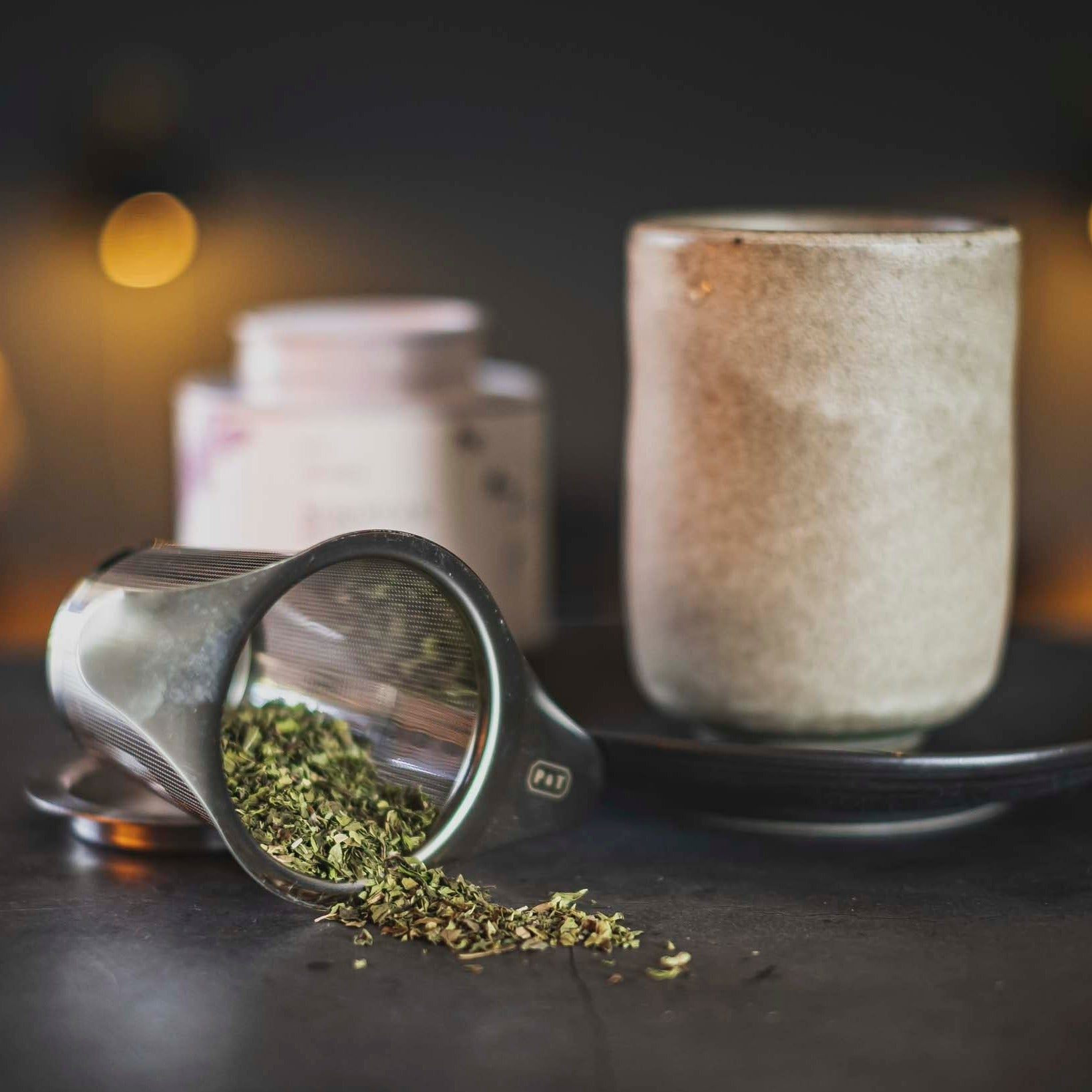 Metall-Tee-Ei und grüne Teeblätter neben einer beige Keramiktasse auf dunkler Untertasse, sanfte Beleuchtung.