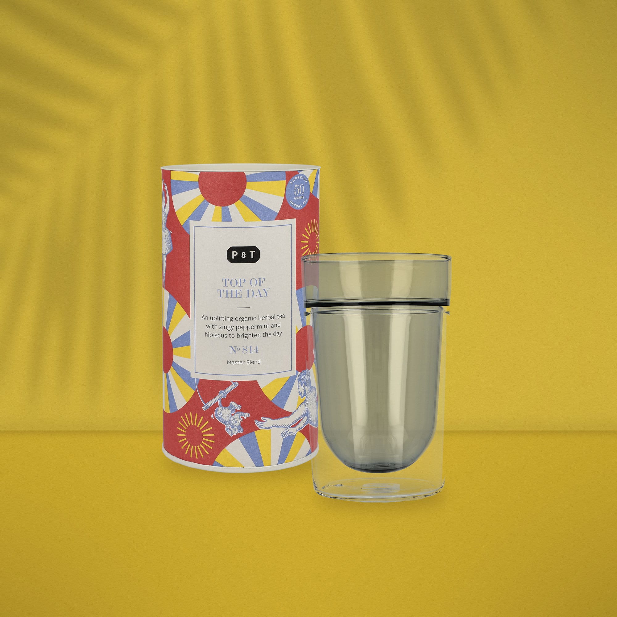 Bunte P & T Teedose 'TOP OF THE DAY' mit Bio-Kräutertee und Doppelwandglas vor gelbem Hintergrund.