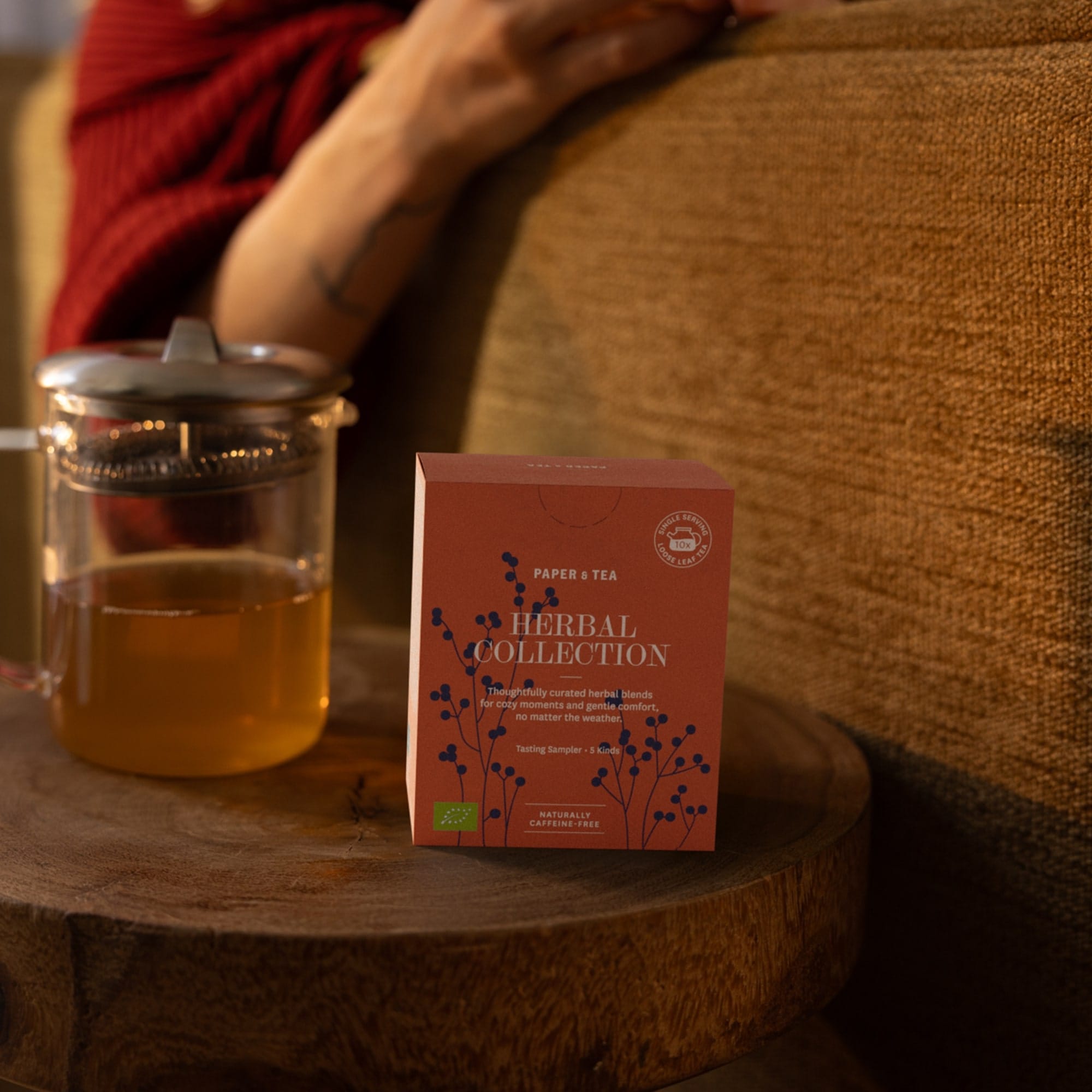 Rote Paper & Tea Teebox mit botanischen Illustrationen und Kräutertee in Glas vor gemütlichem Sofa.