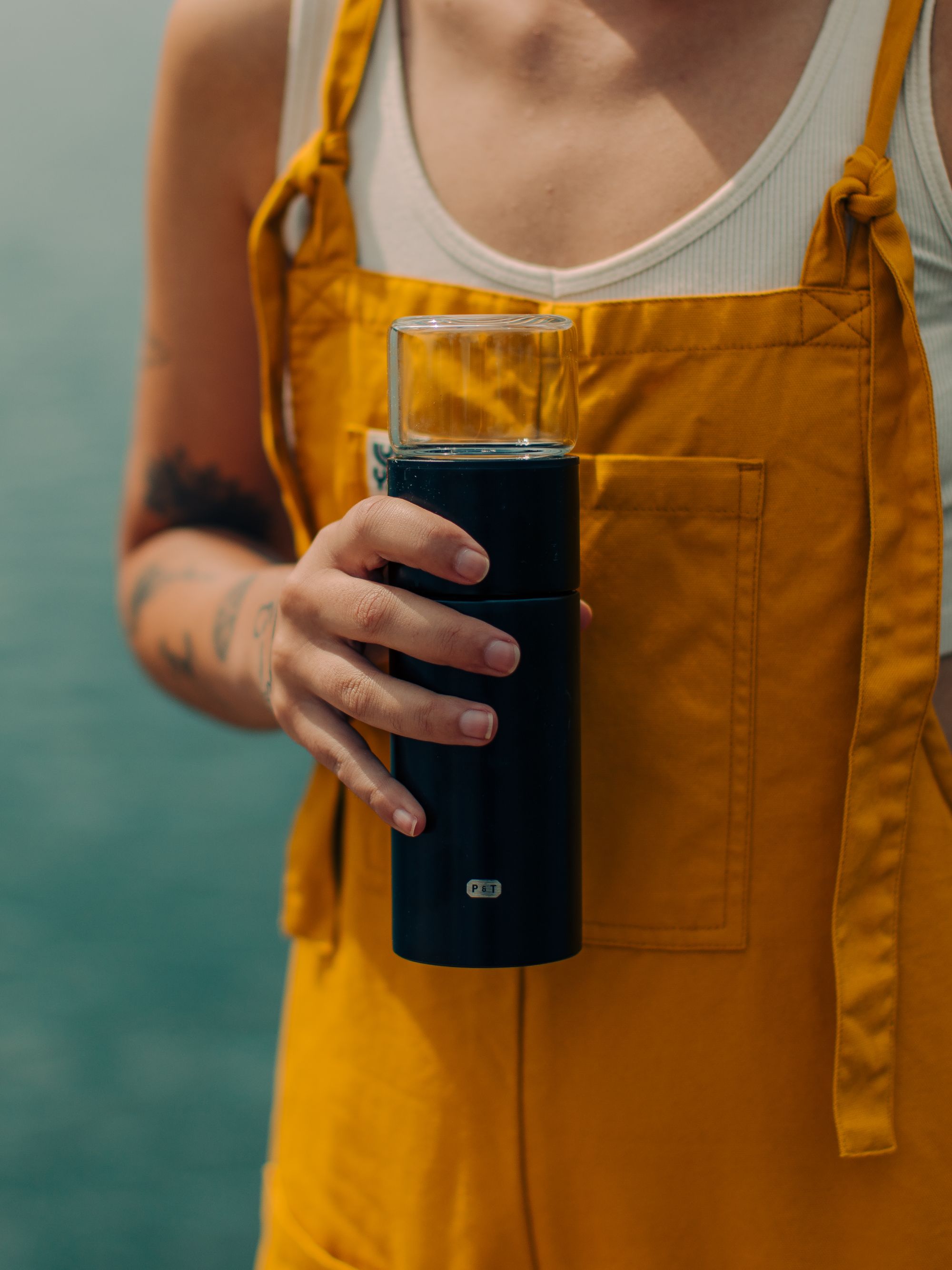 Person in gelbem Latzrock hält eleganten schwarzen Zylinderbehälter mit klarem Deckel und P & T Logo vor einem See.