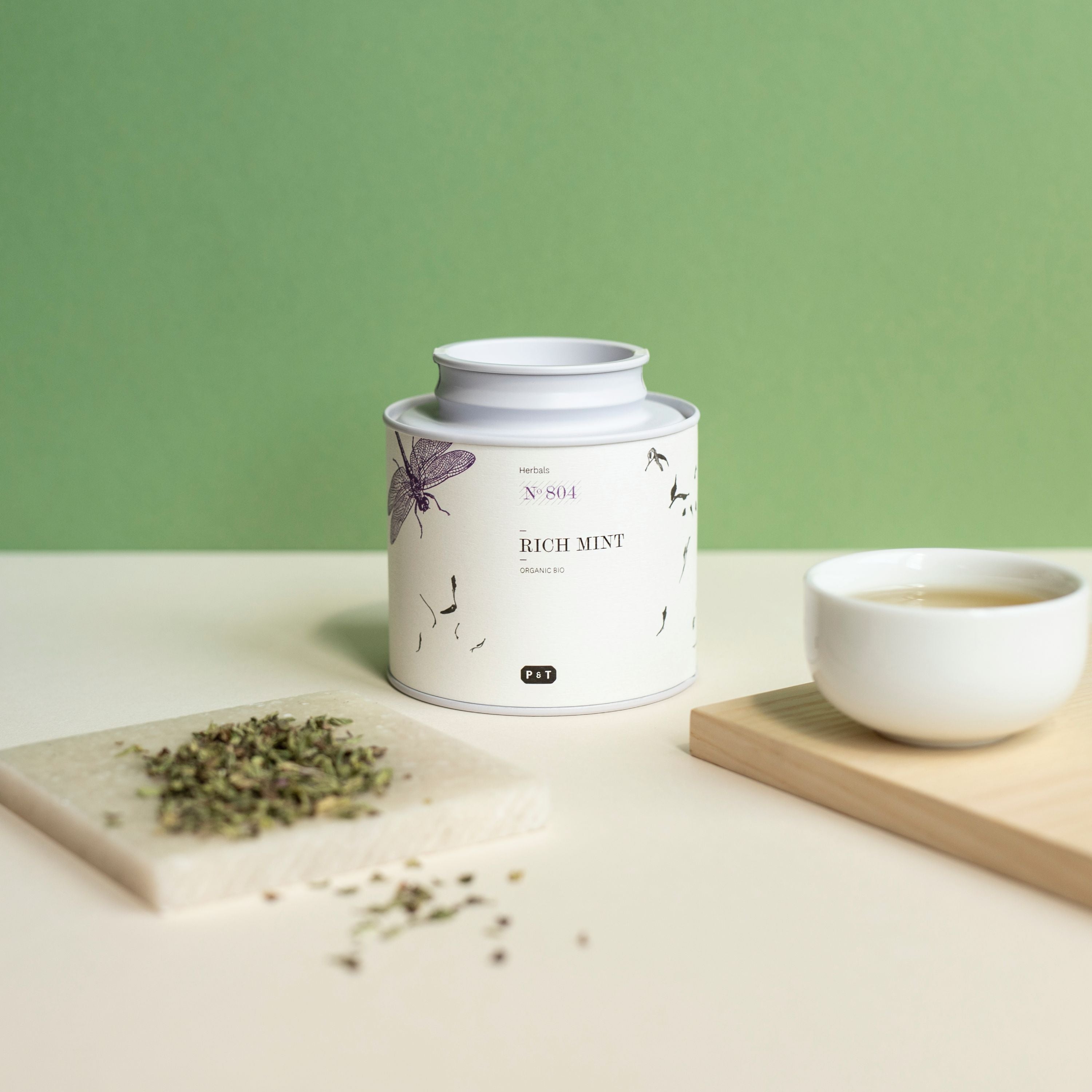Weißer zylindrischer Kräutertee-Behälter mit botanischem Design, Rich Mint No 804, neben Keramiktasse mit hellem Tee.