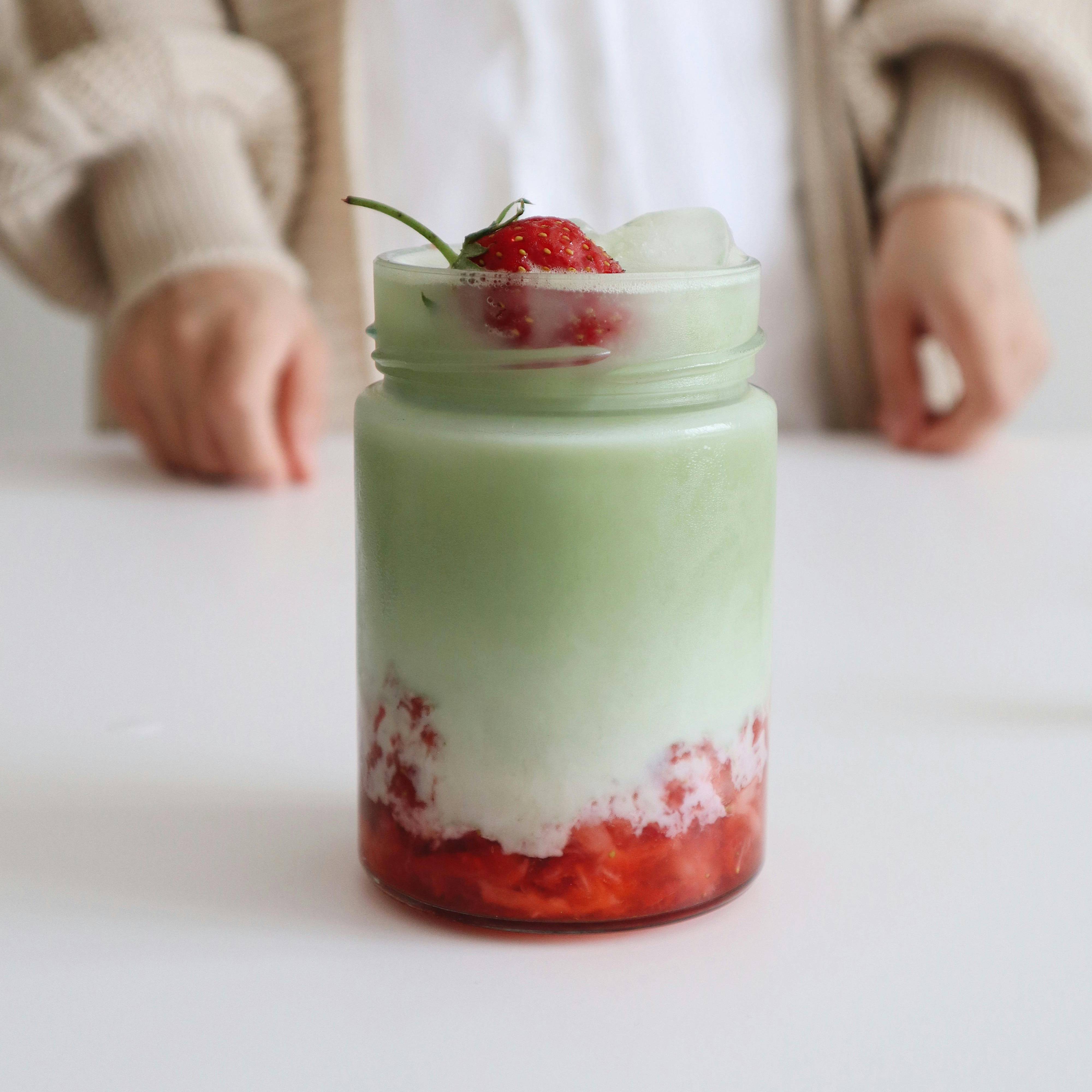 Ein mehrschichtiges Getränk in einem Glas mit rotem Fruchtpüree, grüner Cremeschicht und Erdbeere auf weißem Tisch.