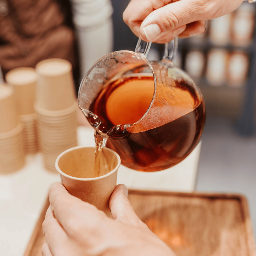 Hand gießt bernsteinfarbenen Tee aus einem Glas in einen braunen Pappbecher vor unscharfem Hintergrund mit Teetassenstapel.