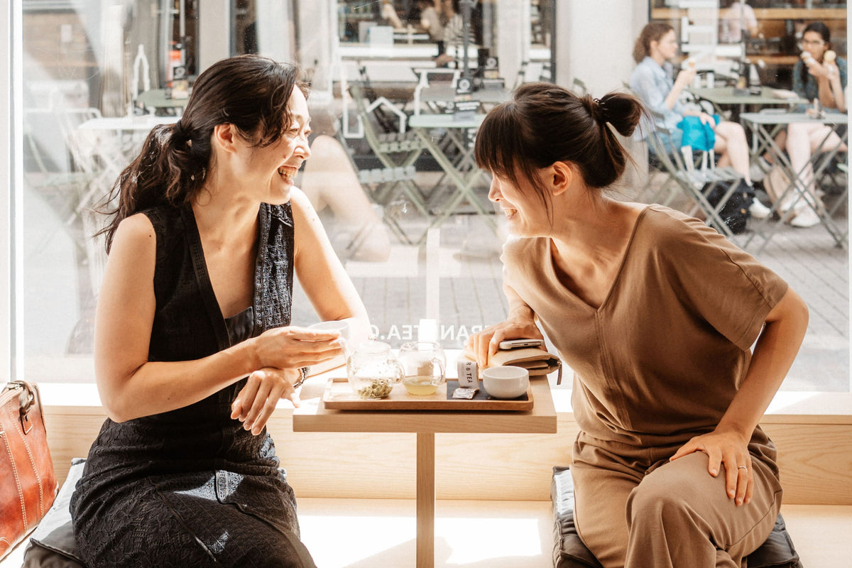 Zwei Frauen in einem hellen Café, trinken Tee aus Glas-Teekannen am Holztisch.