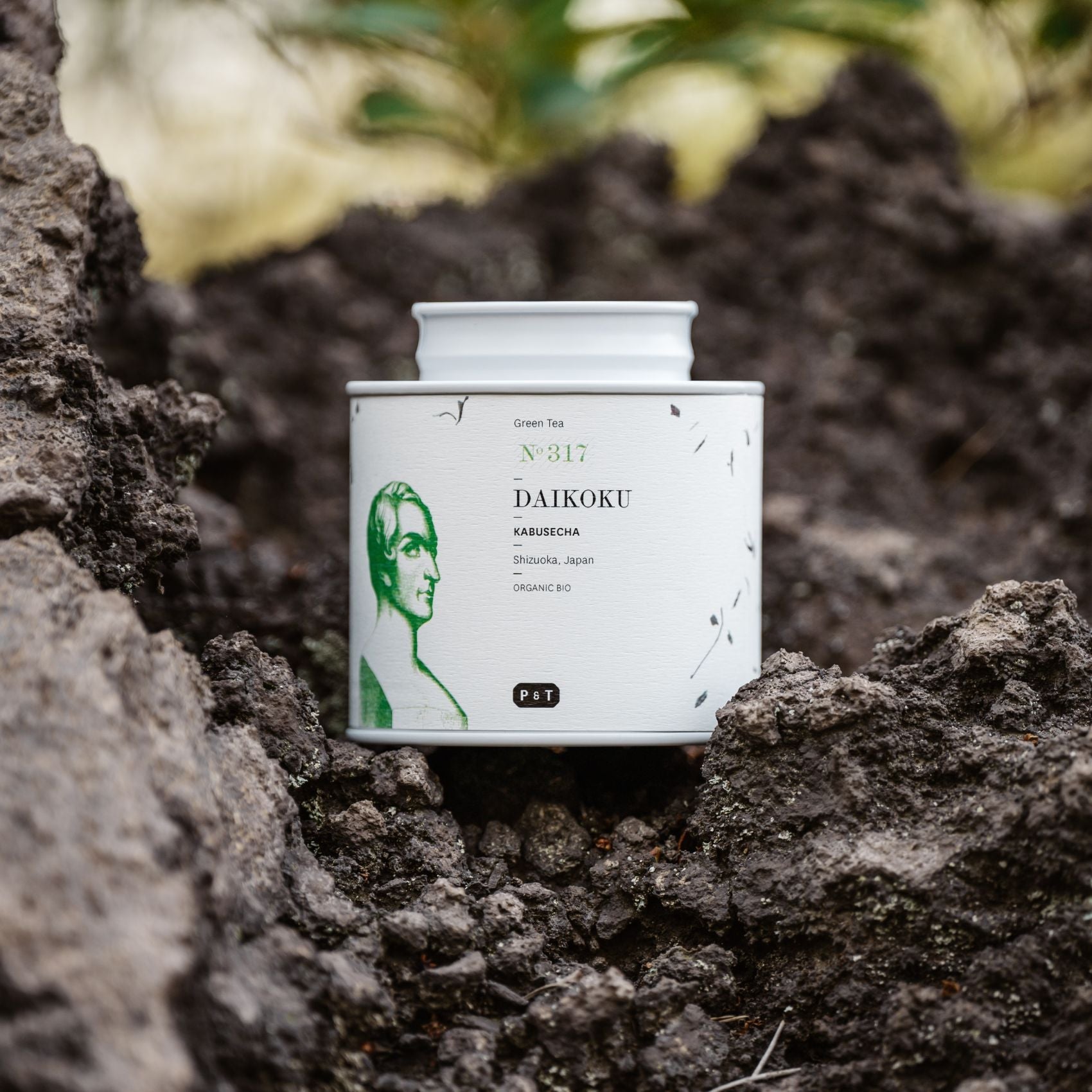 Weiße Teedose mit grünem Porträt und 'DAIKOKU Kabusecha' Text, umgeben von vulkanischen Steinen.