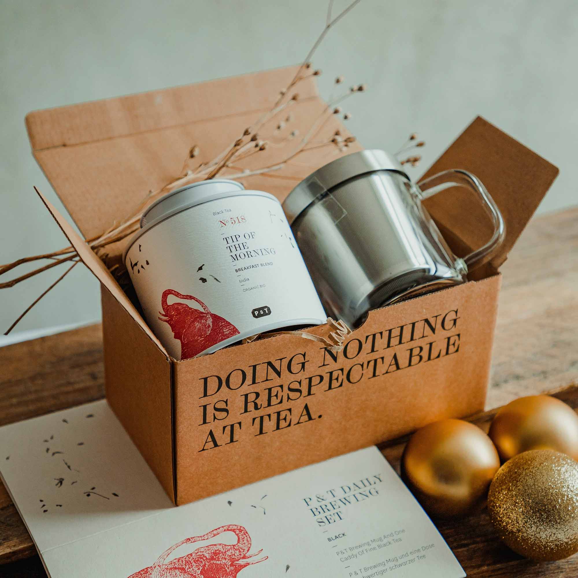 Rustikales P & T Daily Brewing Set mit Bio-Schwarzem Tee, Klarglasbecher, Metall-Infuser und goldenen Kugeln.