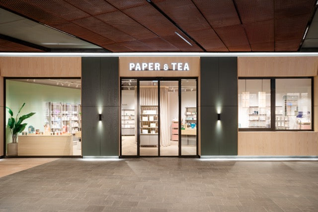 Moderner PAPER & TEA Laden mit Holz- und Glasfront, großen Fenstern und stilvoller Warenpräsentation.