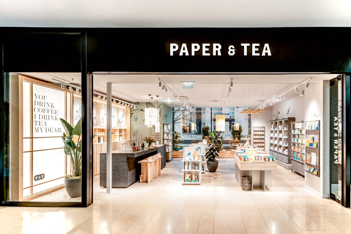 Moderner Laden mit 'PAPER & TEA' Schild, Holzregalen, Pflanzen und großem Leuchtplakat mit Tee-Zitat.