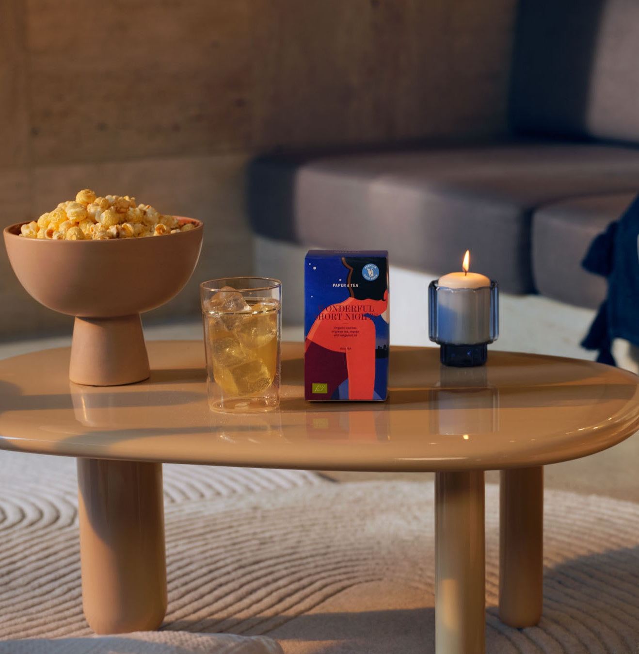 Beiger ovaler Couchtisch mit Papier Teabox 'WONDERFUL SHORT NIGHT', Glas Eistee, Popcornschale und Kerze.