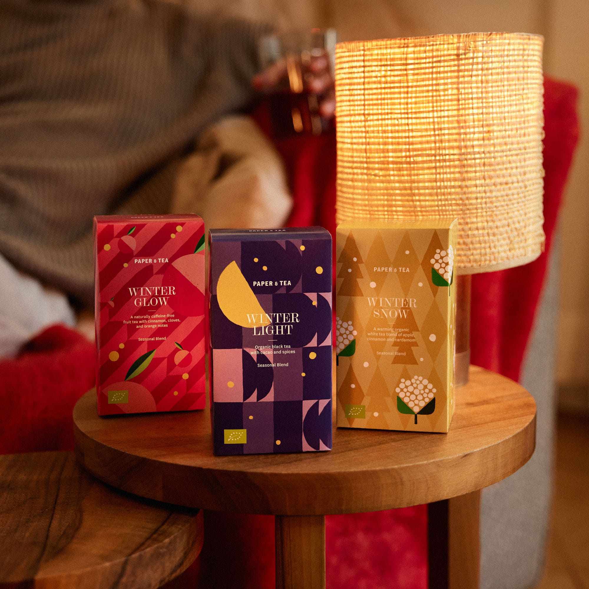 Drei farbenfrohe PAPER & TEA Wintertee-Boxen auf einem runden Holztisch unter warmer Lampe.