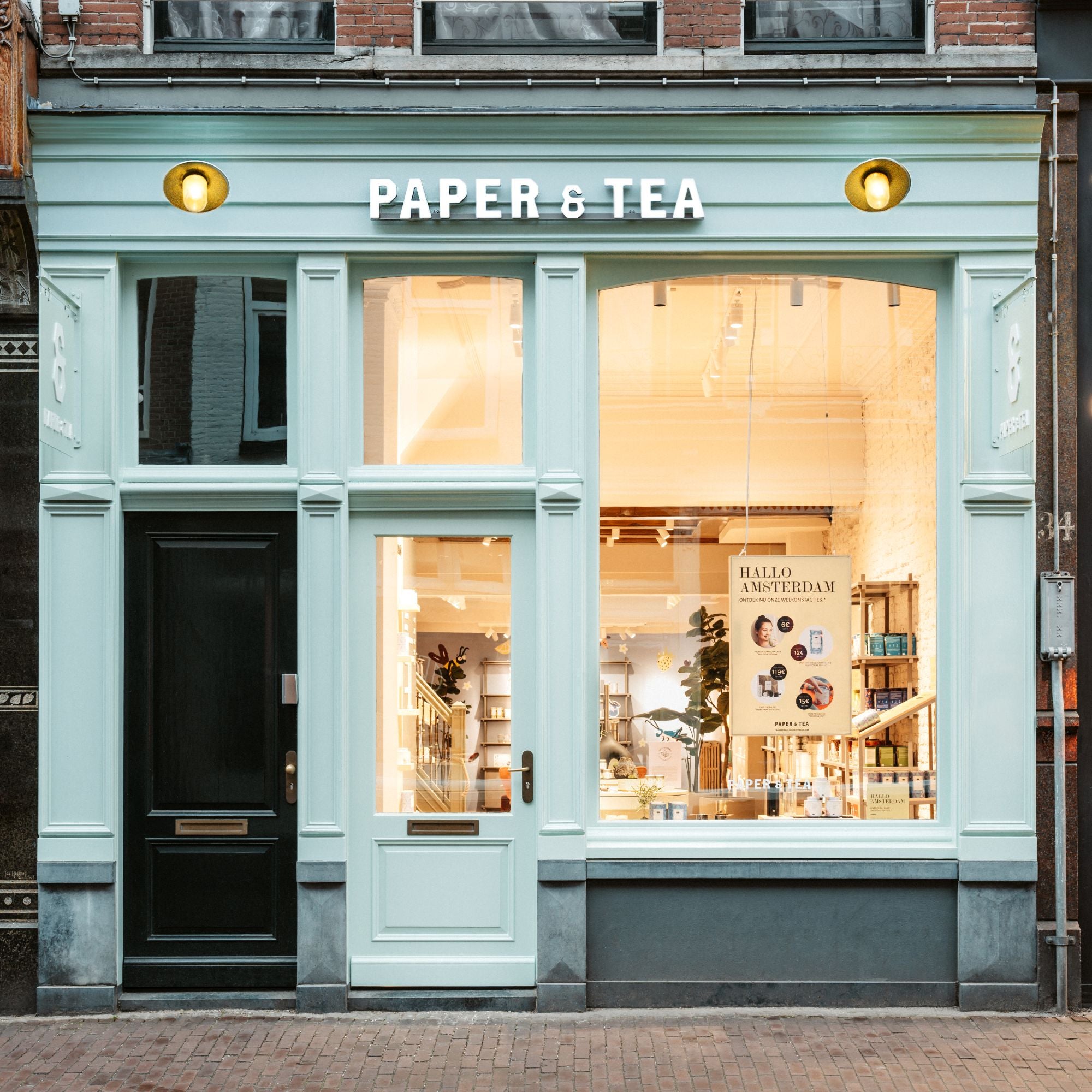Fassade des PAPER & TEA Ladens, pastellgrün, große Fenster, beleuchtetes Logo, 'HALLO AMSTERDAM' Poster