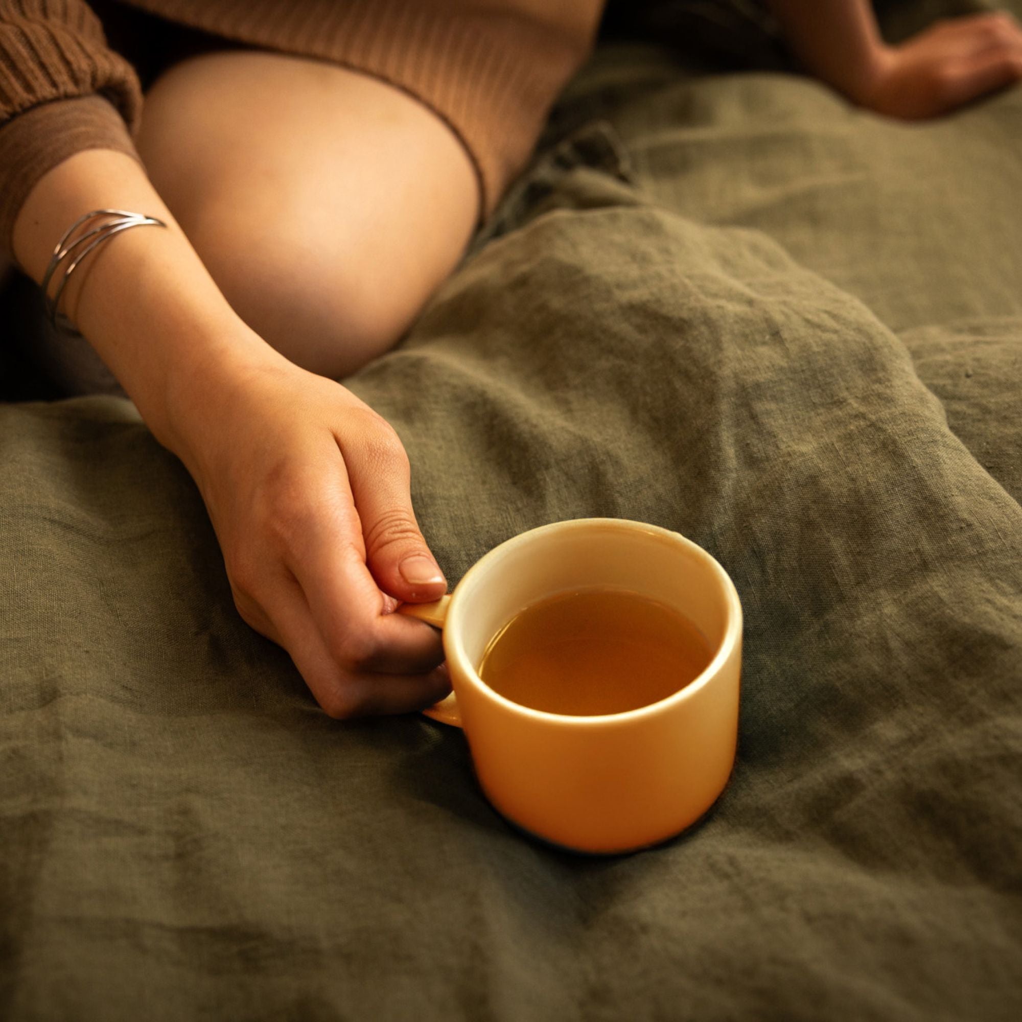Hand im braunen Pullover hält gelbe Keramiktasse mit Tee auf dunklem grünen Stoff.