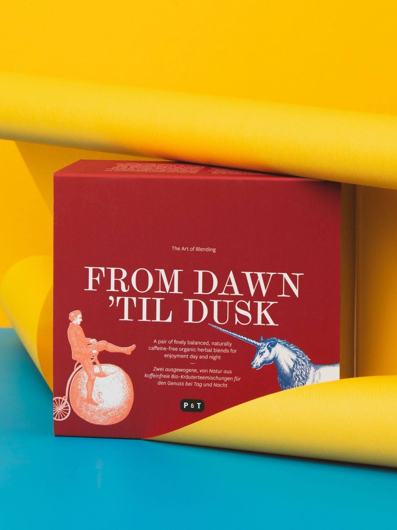 Bio-Kräuterteemischung von Paper & Tea in roter Box mit Vintage-Motiven, gelbe Rollen im Hintergrund.